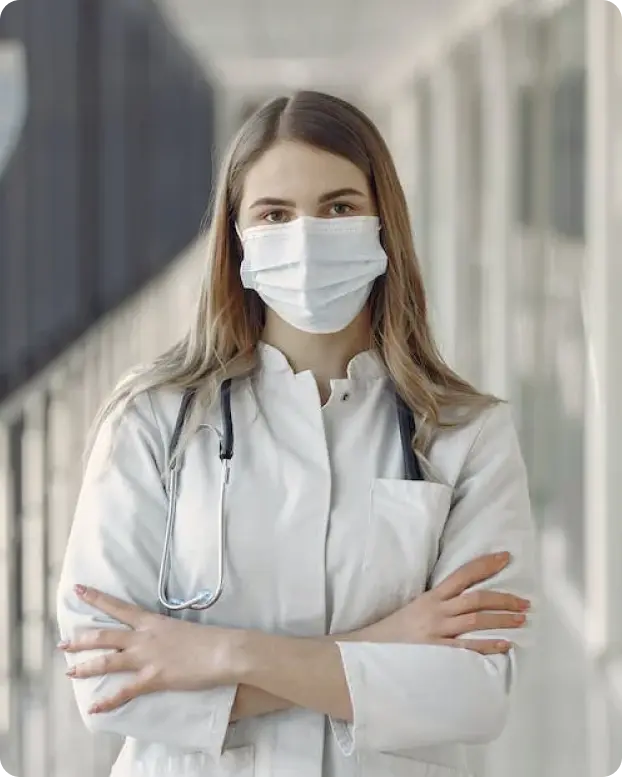 A nurse in a white coat with a stethoscope around their neck stands with arms crossed in a hospital corridor. The background features a modern, well-lit hallway with glass panels, suggesting a professional medical environment.