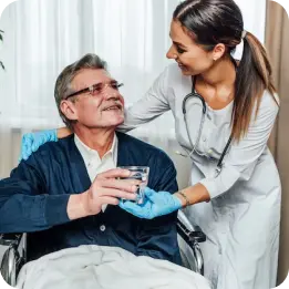 A nurse wearing blue gloves provides elderly care by assisting a senior man in a wheelchair, offering him a glass of water in a comforting hospital room setting.