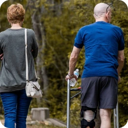 A person assists an individual using a walker outdoors, with the latter wearing a knee brace for support, in a natural setting with trees in the background. The image highlights rehabilitation for orthopedic and sports injuries.