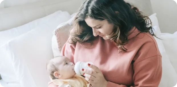 A mother in a pink sweater sitting on a bed, gently feeding her baby with a bottle while looking at the baby lovingly. The baby, dressed in a yellow outfit, gazes up at her.