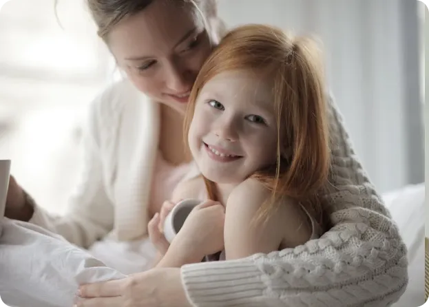 A mother and her young daughter are sitting together. The mother gently embraces her daughter, who has red hair and a bright smile. They both appear happy and content. The daughter is holding a white cup while leaning into her mother’s arms.