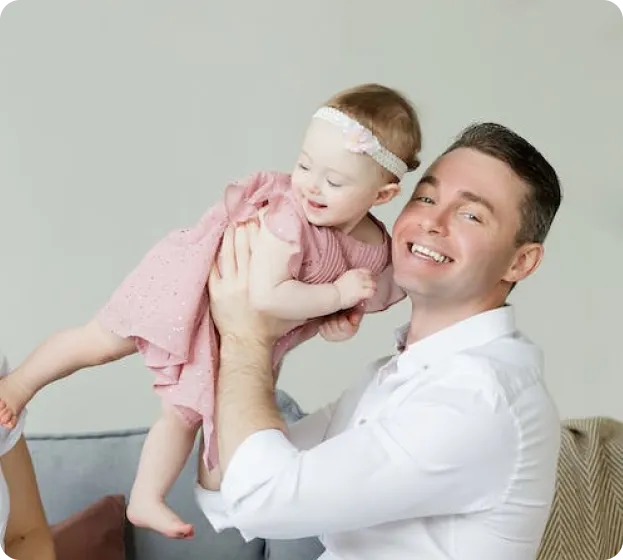 A smiling father in a white shirt holding his baby girl in a pink dress and headband, lifting her playfully while she laughs. A partial view of another person is visible on the left side of the image.