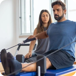 A physiotherapist guides a patient through a neurological rehabilitation exercise on a reformer machine, focusing on strength and mobility in a well-lit room. The image highlights specialized physiotherapy at home for neurological recovery and improved physical function.