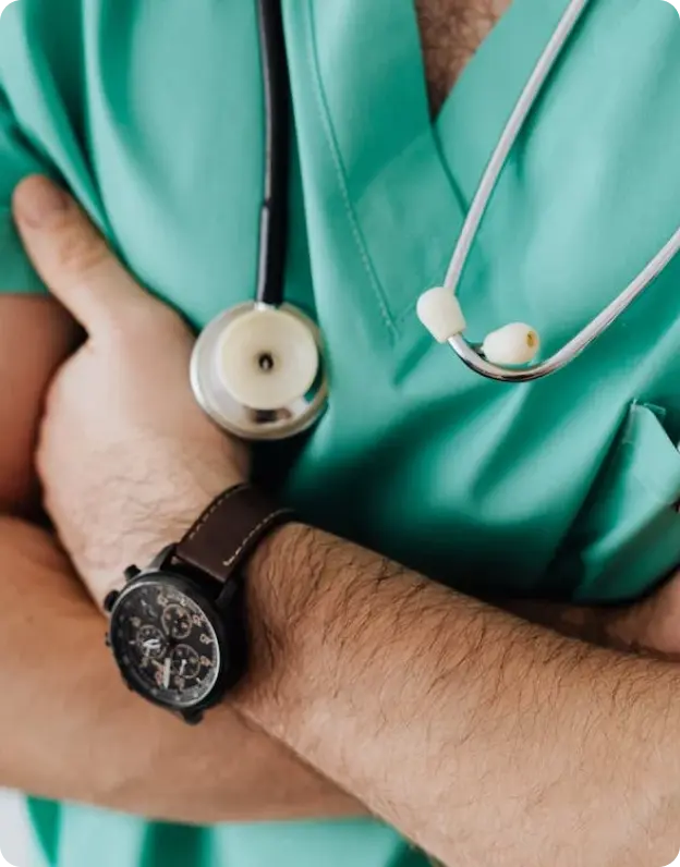 A close-up of a Nurse in a teal uniform with arms crossed, wearing a stethoscope around their neck and a wristwatch. The background is blurred, suggesting a clinical or hospital setting.