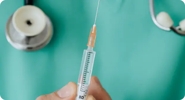 A close-up image of a hand holding a syringe with a needle, filled with a clear liquid, in front of a teal background. In the background, a stethoscope is visible, suggesting a medical setting.