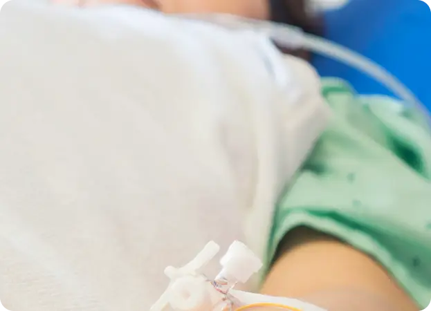 A close-up image of a person's arm with an IV line inserted, connected to a tube. The person is wearing a light green medical gown and is partially covered with a white blanket, indicating a medical or hospital setting.