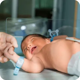 The image shows a newborn baby lying on a hospital bed or incubator, wearing a medical identification bracelet. The baby is reaching out and holding an adult’s finger. This image can effectively represent neonatal care home services, emphasizing professional and compassionate care for newborns in a home setting.