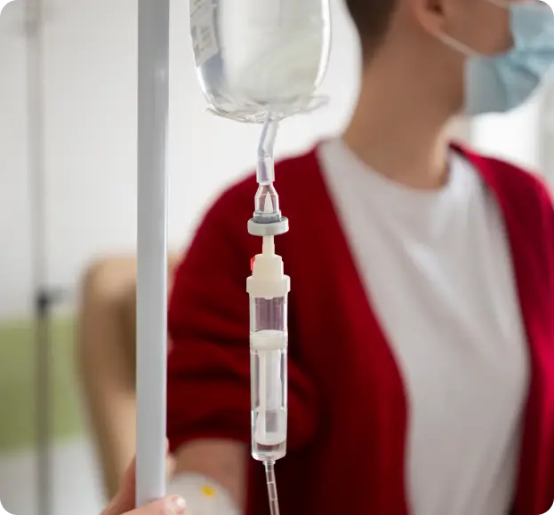 A close-up image of an IV drip setup, showing a bag of fluid hanging on a stand, connected to a tube with a drip chamber. In the background, a person wearing a red cardigan over a white shirt and a face mask is partially visible, suggesting a home environment.
