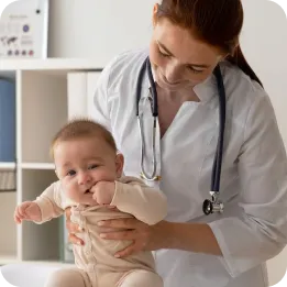 A pediatric healthcare professional wearing a white coat and stethoscope holding a smiling infant during a medical examination. The baby appears healthy and content while being carefully examined in a clinical setting with shelves and medical equipment visible in the background