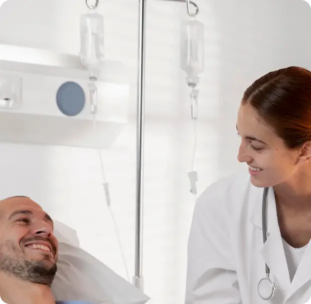 An image of a healthcare professional in a white coat smiling and talking to a patient lying in a hospital bed. The patient is connected to an IV drip with two fluid bags hanging on a stand beside the bed, indicating a medical setting. The background includes a white wall and medical equipment.