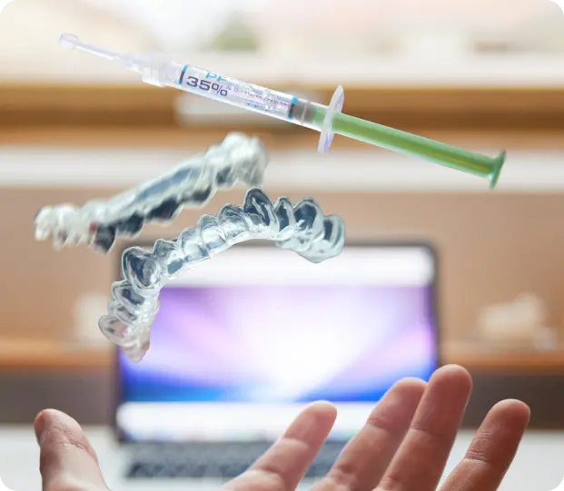 A hand reaching out in the foreground with a blurred laptop in the background, while a syringe and a clear dental aligner appear to be floating in mid-air. The scene represents a blend of medical and dental care technology, featured on the Intelligent Care website, a homecare service in Dubai.