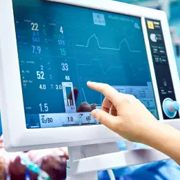 A healthcare professional's hand touches the screen of a medical monitor displaying vital signs, including heart rate, blood pressure, and oxygen levels, in a hospital setting. The image emphasizes vital sign monitoring of patients.