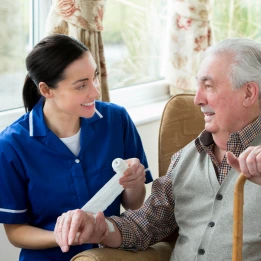 A nurse provides palliative care at home, gently holding an elderly man's hand while showing him a medical item in a cozy living room. The image emphasizes compassionate support for seniors in a home setting.