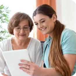 A caregiver in a blue uniform shows a tablet to an elderly woman in a white blouse, both smiling in a cozy room with plants and orange curtains in the background.