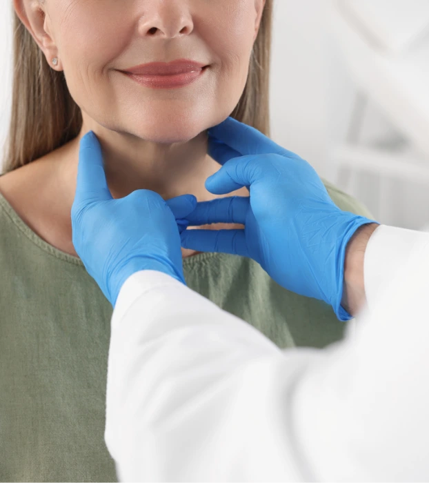 A healthcare professional in a white coat and blue gloves examines a patient's neck, gently palpating the thyroid area. The patient, wearing a green shirt, has long hair and small earrings, with a blurred clinical background.